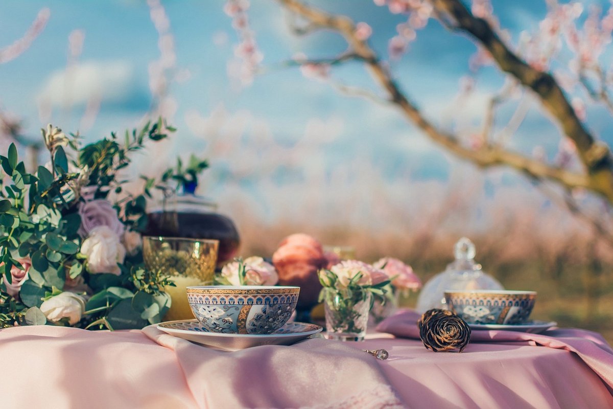 Two patterned porcelain tea cups on saucers on a pink-draped outdoor table, with a glass teapot of dark tea, roses and blossom branches behind